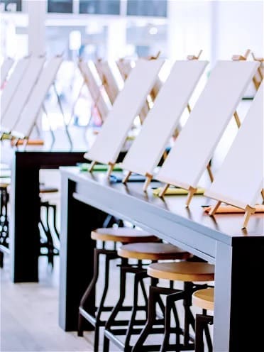 black tables with artist stools below, canvases, easels and paint brushes on top at a Pinot and Picasso Studio